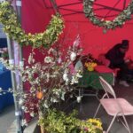 Na obrazku pojawia się stragan z różowym zadaszeniem, na którym są wystawione są dekoracyjne elementy oraz dodatkowe elementy dekoracyjne, które wiszą. Za stołem przykrytym zielonym obrusem siedziba osoby, a obok znajduje się kosz znajdujący się w magazynie.