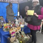 Na obrazku kobieta trzyma tabliczkę z napisem „Rada Rodziców” przy straganie, na której są wyeksponowane, są odpowiedzialne za robione ozdoby, w tym figurki zwierząt i określenia kwiatowe. Dwie kobiety zastępcze za stołem pod pasowanym zadaszeniem.