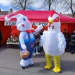 Dwaj ludzie przebrani za królika i kurczaka na zewnątrz targowiska przed czerwonym namiotem. Królik trzyma koszyk i gestykuluje w stronę kurczaka, gdy w pobliżu znajduje się inna osoba.