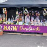 Na obrazku pojawia się stoisko, za które stoi osiem osób używających uszu królika. Na stoły wyeksponowane są proste wypieki, wieńce oraz doniczkowe kwiaty, a całość jest oznaczona banerem z napisem "KGW Porębianki".