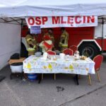 Na obrazku widoczni są strażacy znajdujący się pod namiotem oznaczonym napisem „OSP Mlecin”. Siedzą przy udekorowanym stole z przekąskami i napojami, a w tle stoi czerwony pojazd strażacki.