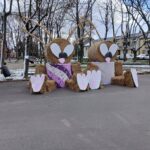 Na obrazku znajdują się dwie duże rzeźby królików wykonane z belsiana, które mają pomalowane oczy, uszy i łapy. W tle widać drzewa z rzadkimi składnikami oraz osobą, która pochodzi z lewej strony.