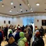 Grupa ludzi w kolorowych kurtkach w sali wystawowej z rzeźbami. Jasne ściany ozdobione popiersiami i obrazami. Nowoczesne oświetlenie sufitowe.