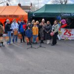 Na rynku występuje grupa osób utworzonych przed pomarańczowymi i zielonymi namiotami, z mikrofonami występującymi na pierwszym planie. Niektóre trzymają torby z prezentami, a po prawej stronie jest baner z napisem „25 lat”.