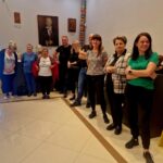 Grupa ludzi stoi w jasnym pokoju z popiersiami na ciemnych meblach. Ściany zdobią obrazy i girlanda z kwiatów. Podłoga jest kremowa, a obok stoi żółty znak ostrzegawczy.