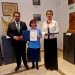 Trzy osoby stoją w galerii z dziełami sztuki. Środkowa osoba trzyma dyplom, po prawej są rzeźby i obrazy. Tło jest jasne i neutralne.