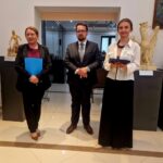 Trzy osoby w eleganckich strojach stoją w galerii sztuki. Tło to jasne ściany i rzeźby. Dominują kolory czarny, biały i beżowy.