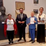 Cztery osoby stoją w muzeum. Kobieta w niebieskim trzyma dyplom, obok rzeźba popiersia. Tło jest jasne z obrazami.