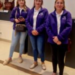 Trzy kobiety w fioletowych kurtkach stoją na jasnym tle. Ubrania mają logotyp KGW Porębianie. Za nimi widoczne są ciemne i brązowe elementy dekoracyjne.