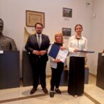 Trzy osoby stoją w eleganckim pomieszczeniu galerii pełnej sztuki. Mężczyzna w garniturze i dwie kobiety trzymają dyplomy. W tle widoczne rzeźby i obrazy na jasnych ścianach.