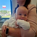 492074404_701957188846472_2454649392822449072_n Na zdjęciu kobieta w różowej marynarce trzyma uśmiechnięte niemowlę w białym ubranku. W tle przez okno widać polską flagę i zielone drzewa. Obok na stole stoi bukiet kwiatów.