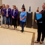 Grupa ludzi stoi na tle małych posągów w galerii. Kobiety ubrane w fioletowe i niebieskie koszulki. Elegancka kobieta z niebieską teczką.