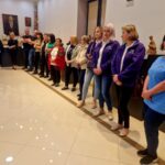 Grupa ludzi stoi w galerii sztuki, na jasnej podłodze, w tle widać popiersia i obrazy na ścianie. Kilka osób nosi fioletowe bluzy, jedna ma niebieską koszulkę. Pomieszczenie jest dobrze oświetlone.