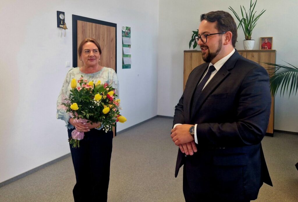 Na zdjęciu kobieta z bukietem żółtych i różowych kwiatów, obok mężczyzna w garniturze. Tło to jasne biuro z roślinami. Na szafce stoi zdjęcie.