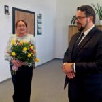 492298395_701957128846478_3570740181546120971_n Na zdjęciu kobieta z bukietem żółtych i różowych kwiatów, obok mężczyzna w garniturze. Tło to jasne biuro z roślinami. Na szafce stoi zdjęcie.