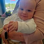 493085378_701956965513161_7285021813873444790_n Na zdjęciu jest uśmiechnięte niemowlę na rękach dorosłego. W tle widać flagę Polski na maszcie i niebieskie niebo. Dziecko ma na sobie białe ubranko.