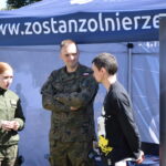 Na zdjęciu są trzy osoby w rozmowie, jedna w mundurze, na tle stoiska rekrutacyjnego Wojska Polskiego. Tło jest niebieskie z napisem zostanzolnierze. Widać jasne słońce i cienie wskazujące na dzień.