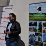 Mężczyzna w czarnej kurtce stoi obok dwóch banerów. Na jednym widnieje napis Mazowsze, a drugi promuje Gminę Dobre z krajobrazami. Tło jest jasnoszare, a scena oświetlona słońcem.