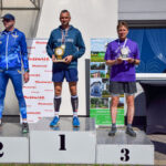 Trzech mężczyzn na podium z trofeami, w sportowych strojach. Tło to biały budynek i plakaty. Słońce jasno świeci.