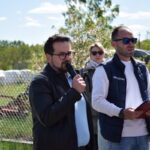 Mężczyzna w czarnej kurtce trzyma mikrofon, przemawiając na świeżym powietrzu. Obok niego stoją dwie osoby. W tle zielone drzewa i ogrodzenie.