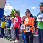 Grupa dzieci w kolorowych strojach stoi na starcie biegu, ubranych w numery startowe. Na tle widać niebieski balon startowy i jasne niebo. Dookoła stoją kibice i zieleń, co sugeruje, że wydarzenie odbywa się na świeżym pow