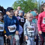 Dzieci w kolorowych sportowych strojach z numerami startowymi gotowe do biegu. Tło to zielone drzewa, jasne niebo. Twarze są skupione i uśmiechnięte.