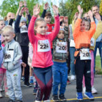 Grupa dzieci w kolorowych strojach z numerami startowymi podnosi ręce na biegu. W tle zielone drzewa i niebo. Wydarzenie sportowe na świeżym powietrzu.