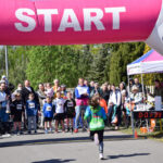 Start biegu dziecięcego, kolorowe ubrania, tłum kibiców. Nad biegaczami różowy balon z napisem START. W tle zielone drzewa.