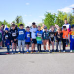 Grupa dzieci stoi na linii startu wyścigu. W tle widoczni są kibice oraz zielone drzewa pod błękitnym niebem. Obok jest zegar startowy z czerwoną cyfrą 0:00.