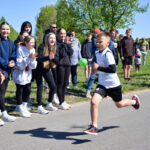 Na zdjęciu jest chłopiec biegnący po asfaltowej drodze w białej koszulce. W tle ludzie kibicują, niebo jest niebieskie. Drzewa i trawa dopełniają zielone tło.
