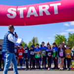 Grupa dzieci stoi na starcie wyścigu pod różowym łukiem z napisem START. Mężczyzna w niebieskiej kamizelce trzyma mikrofon. Tło to jasne niebo i drzewa.