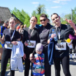 Grupa osób w strojach sportowych po biegu, na tle drzew i budynku. Uśmiechają się, trzymając numery startowe i butelki wody. Słoneczny dzień.