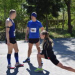 Trzech biegaczy w sportowych ubraniach rozciąga się na tle zielonych drzew. Przeważają niebieskie i czarne kolory. Scena odbywa się na brukowanej ścieżce.