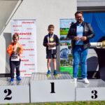 Podium z trzema osobami, z czego dwóch chłopców z medalami na miejscach 1 i 2. Tło białe z czerwonymi napisami Mazowsze. Po prawej stronie stolik z trofeami obok mężczyzny.