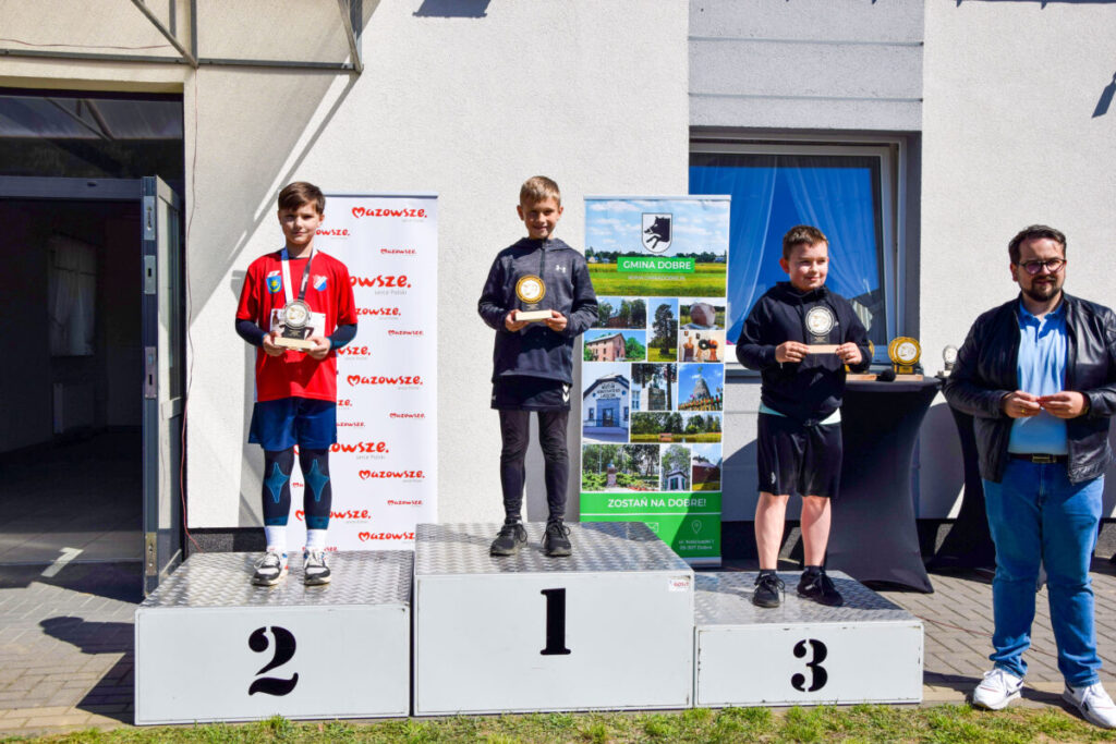 Na zdjęciu trzech chłopców stoi na podium z medalami. Tło jest białe z plakatami i banerem. Są słoneczne i jasne barwy.