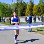 Zawodnik w niebieskim stroju przekracza linię mety maratonu na asfaltowej drodze. W tle zielone drzewa i kibice. Napis Gratulacje na taśmie.