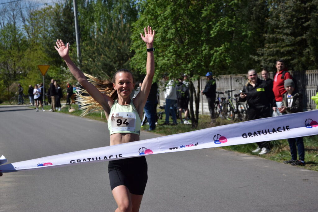 Kobieta przekracza metę maratonu z uniesionymi rękami, uśmiechnięta. Ma na sobie zieloną koszulkę i czarne spodenki. W tle widoczna jest zielona roślinność i kibice.