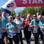 Grupa uczestników biegu stoi uśmiechnięta z medalami na tle dmuchanej bramy startowej. Noszą kolorowe stroje sportowe. W tle zielone drzewa i niebo.