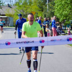 Mężczyzna w limonkowej koszulce przekracza linię mety. Dookoła kibice i uczestnicy, tło z zielonymi drzewami.