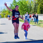 Na drodze kobieta z numerem startowym i dziecko w różowej kurtce biegną, uśmiechając się. W tle zielone drzewa i kibice. Słoneczny dzień.