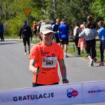 Kobieta w pomarańczowej koszulce przekracza linię mety biegu. Uśmiecha się, tło stanowią drzewa i widzowie. Na pierwszym planie widać biały baner z napisem GRATULACJE.
