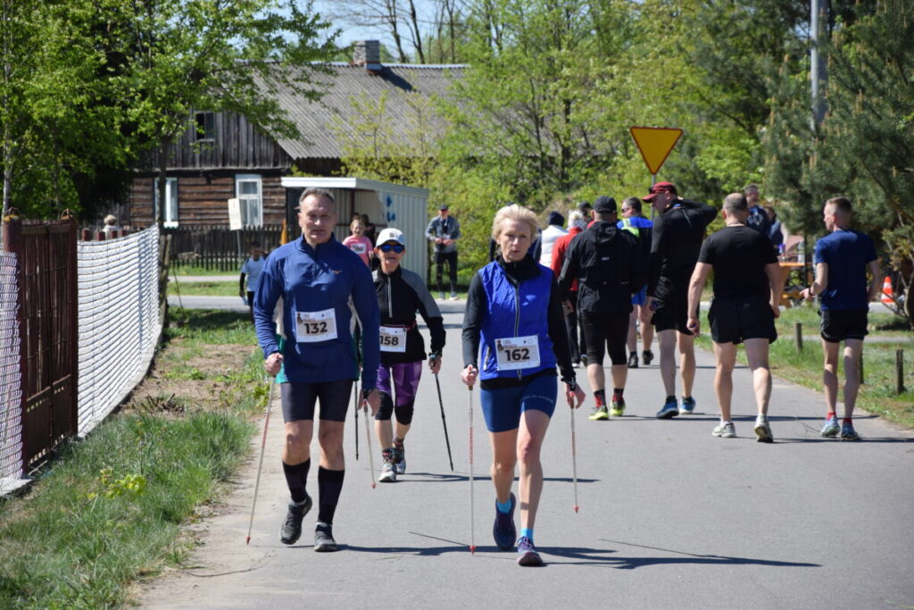 Na drodze odbywa się marsz Nordic Walking. Uczestnicy noszą numery startowe i stroje sportowe. W tle widać zielone drzewa i wiejską chatę.