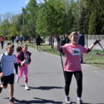 Na zdjęciu ludzie biegną po asfalcie, wśród zieleni drzew. Osoba w różowej koszulce z kijkami marszowymi uśmiecha się. W tle są inni uczestnicy i widzowie.