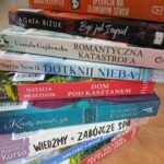 Stos książek na stole z kolorowymi okładkami, w tle regały z książkami. Pomarańcz, czerń, biel i różne odcienie niebieskiego. Napisy na grzbietach wskazują tytuły i autorów.