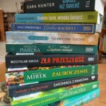 Stos książek na drewnianym stole w bibliotece. Okładki w różnych kolorach, od zielonego po niebieski. W tle rozmyte regały z książkami.