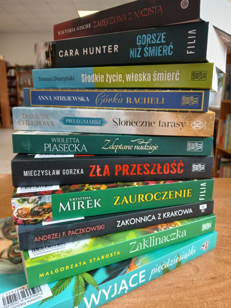 Stos książek na drewnianym stole w bibliotece. Okładki w różnych kolorach, od zielonego po niebieski. W tle rozmyte regały z książkami.