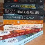 Stos książek o różnobarwnych grzbietach leży na drewnianym stole. Każda książka ma inny kolor okładki i widoczny jest tytuł. Tło jest niewyraźne z półkami książek.