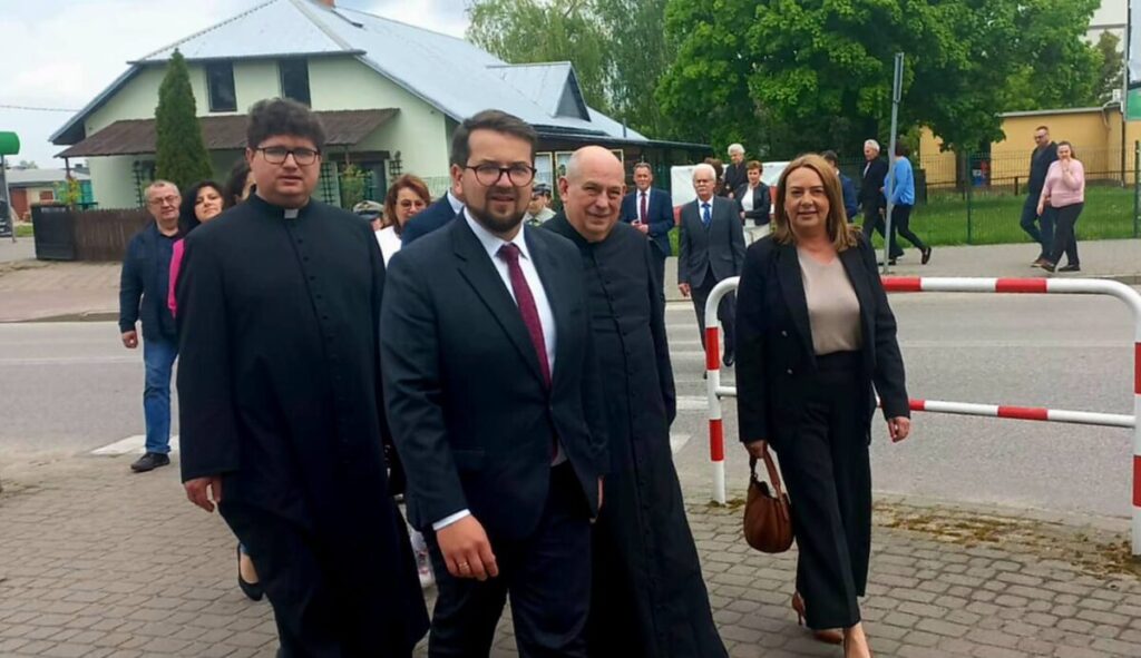 Grupa osób w formalnych strojach idzie po chodniku. W tle zielone drzewa i biały budynek. Pogodny dzień, niebo lekko zachmurzone.