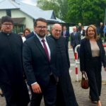 Grupa osób w formalnych strojach idzie po chodniku. W tle zielone drzewa i biały budynek. Pogodny dzień, niebo lekko zachmurzone.