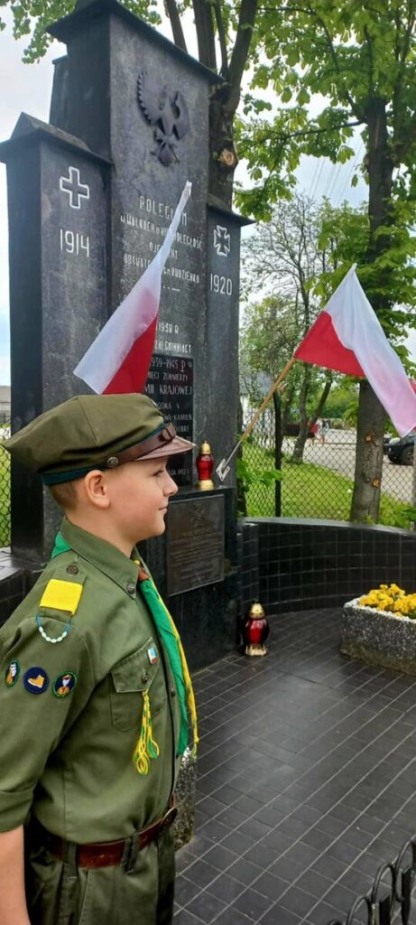 Chłopiec w harcerskim mundurze stoi przed pomnikiem z polskimi flagami. Monument jest ciemny, z białym napisem, tło zielone. Obok znajdują się znicze i kwiaty.